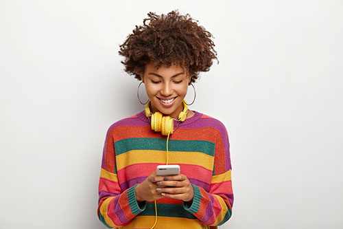 Smiling young woman with curly hair wearing a colourful striped sweater and yellow headphones, looking at her smartphone in a relaxed and casual pose against a plain background.
