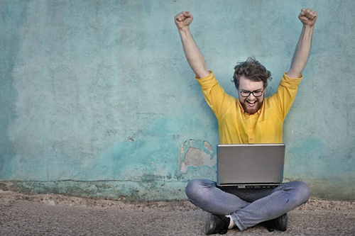 A man sitting on the ground with a laptop, arms raised in celebration or excitement.